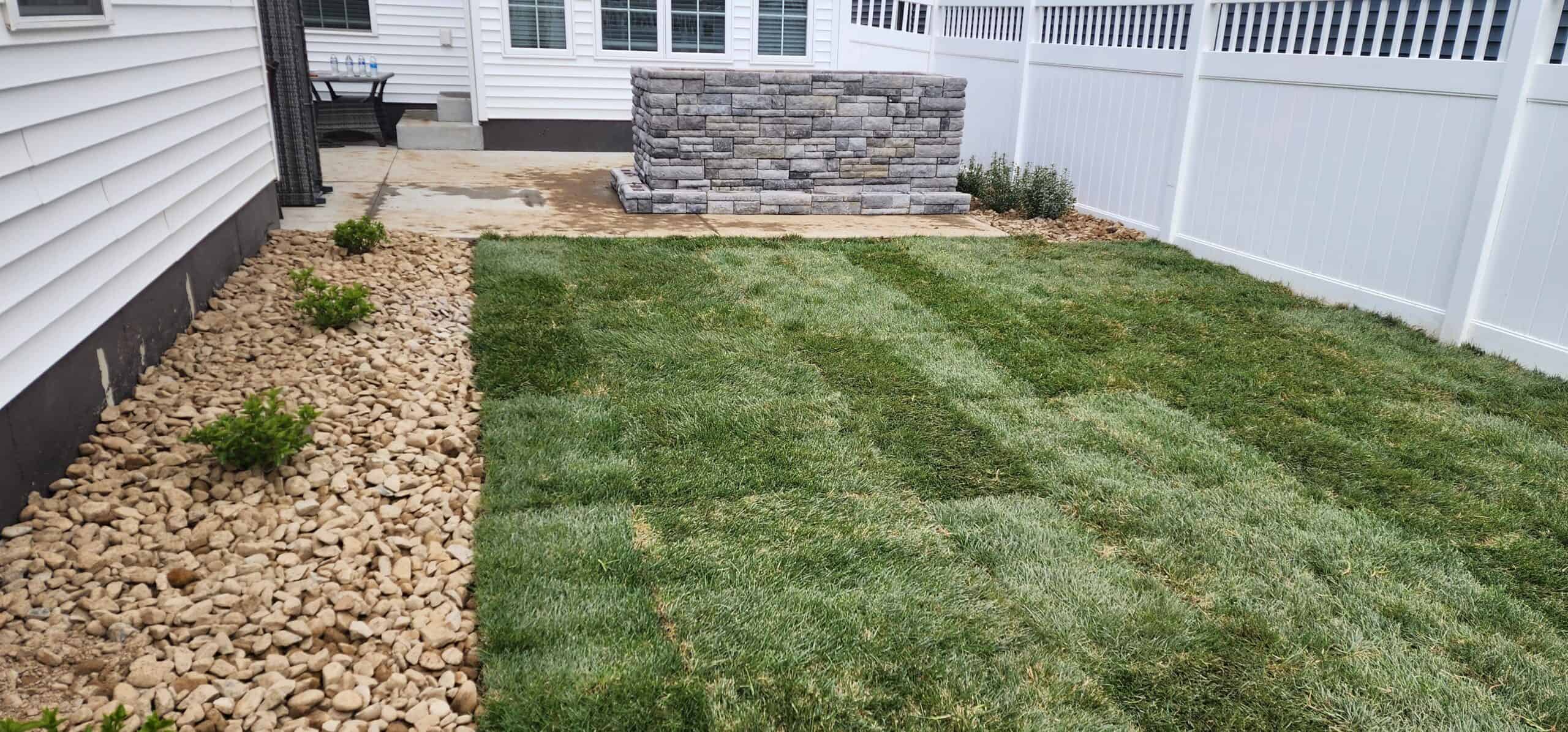a stone firepit on a small backyard patio surrounded by a white privacy fence and a landscaped lawn